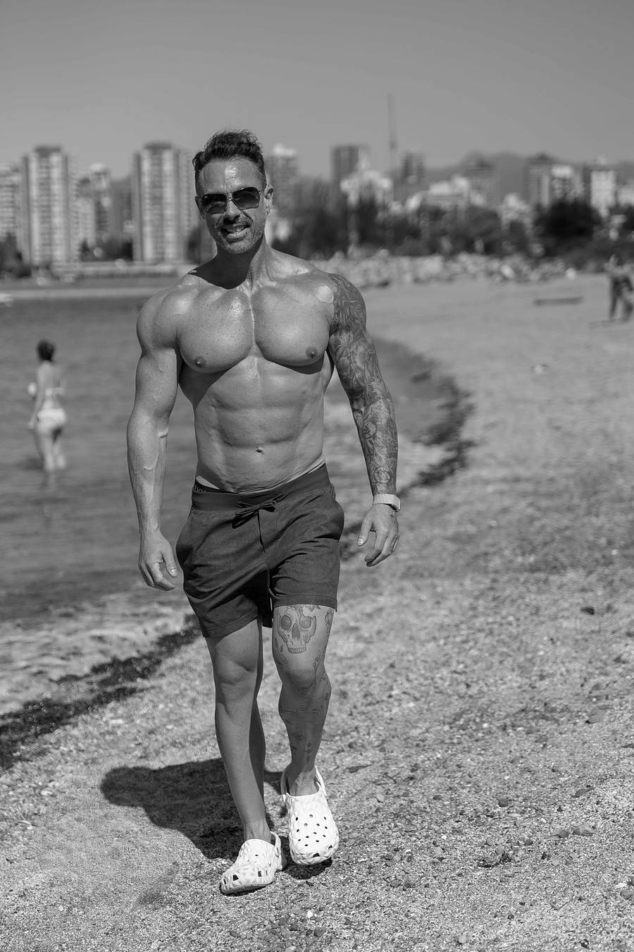 Black and white photo of David Turnbull in sunglasses and shorts walking along a Vancouver beach with the city skyline behind him